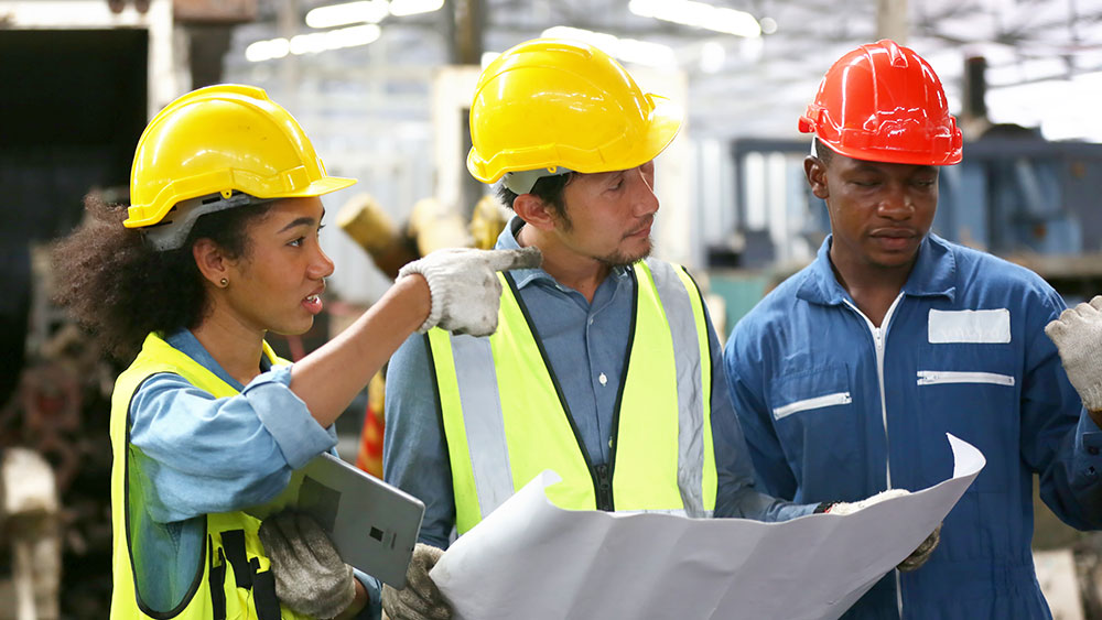 Three engineers in hard hats and safety vests discuss a project together viewing blueprints and a tablet