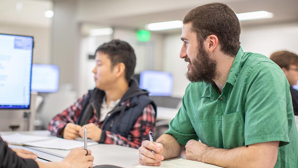 A male student in a green shirt holds a pen as he looks at a monitor. Another student is seen in the background.