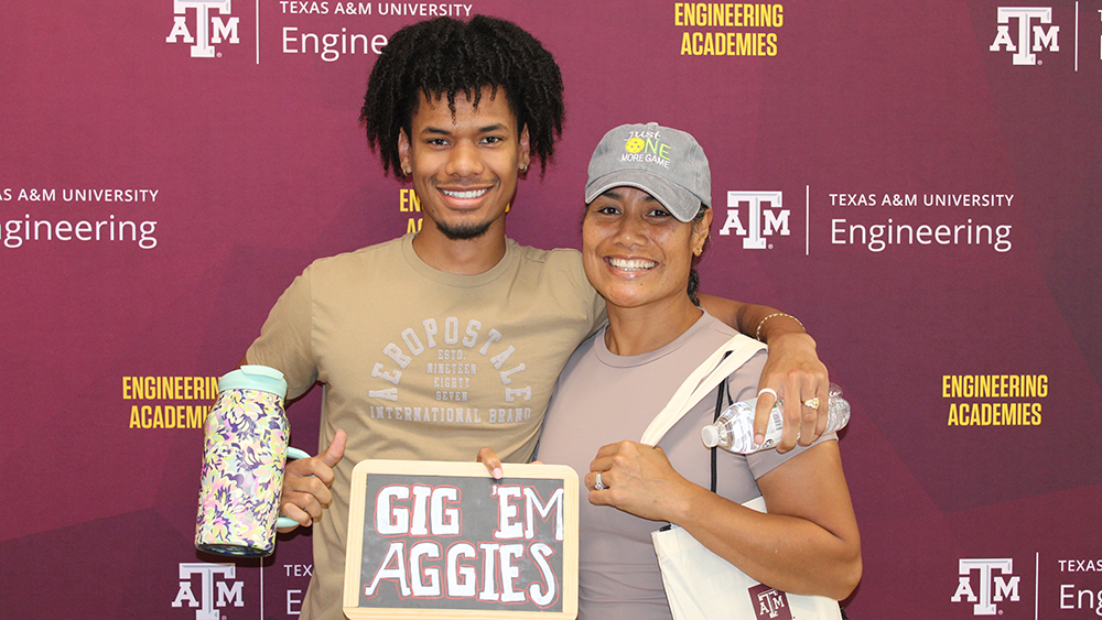 A young man and woman smiling in front of a Texas A&amp;M University Engineering Academies backdrop. The man holds a small "Gig 'Em Aggies" sign while the woman holds a water bottle and wears a hat.