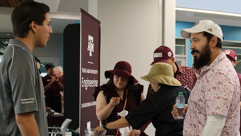 Male wearing a grey polo shirt talks to another man wearing a patterned maroon and white polo shirt. A group of three people take items off a table behind the two men. 