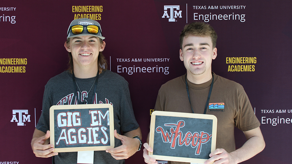 Two young men posing with chalkboard signs in front of a maroon Texas A&amp;M Engineering Academies backdrop. One sign reads "Gig 'Em Aggies" and the other says "Whoop."