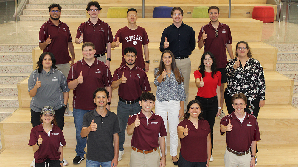 A group of students wearing maroon, white and black clothing stand on a set of stairs smiling at the camera giving thumbs up. 