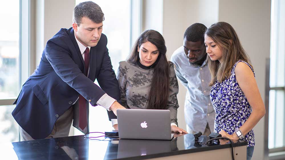 two men and two women gathered around to look at a computer laptop screen.