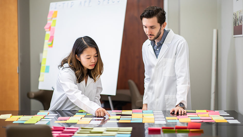 A male and female in lab coats standing in front of a table full of post-it notes