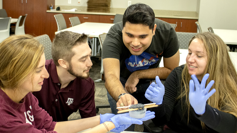 Four students gather around a table. One is holding a 3D model in molding liquid. They are discussing their project.