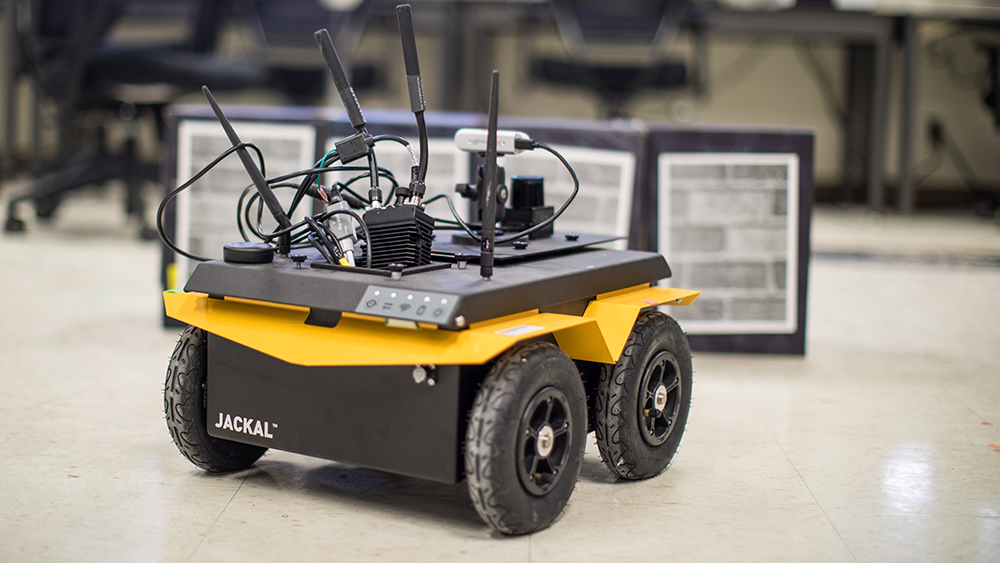 A small robotic vehicle with a box-shaped base sporting four wheels and a tray-like top supporting a flat array with several thick antennas and wired components. 