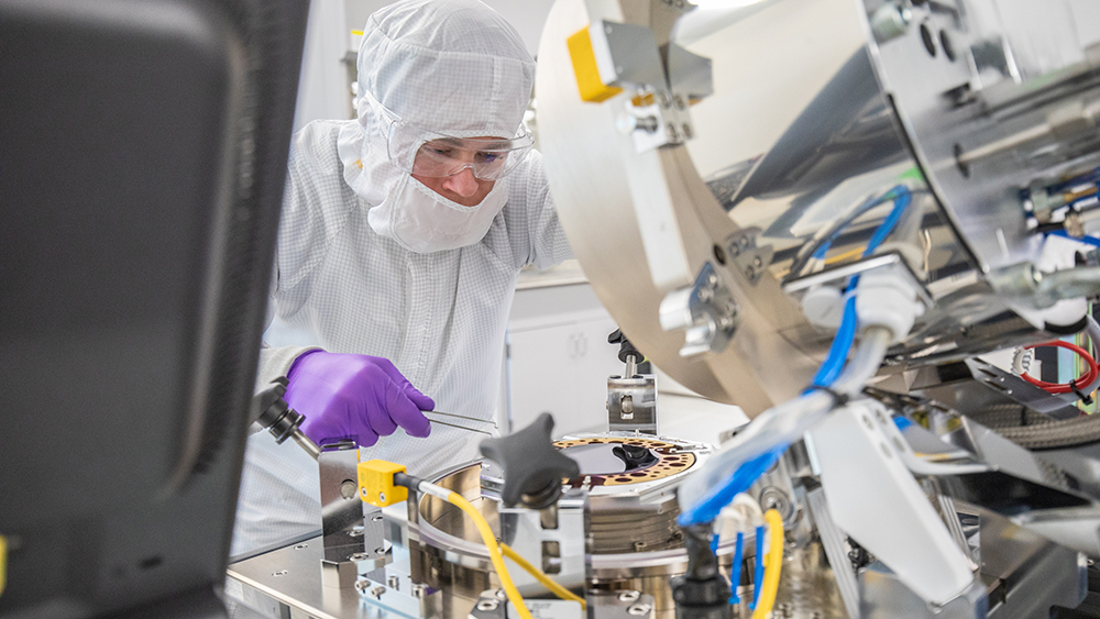 A person in protective lab attire and goggles inspects a small device on a disc-like table surrounded by much larger and more complex equipment.