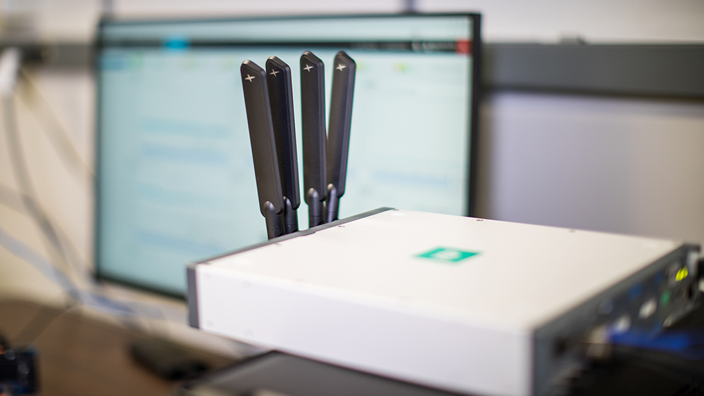 A box-like modem with four thick antennae is in the foreground, with a large computer screen in the background.