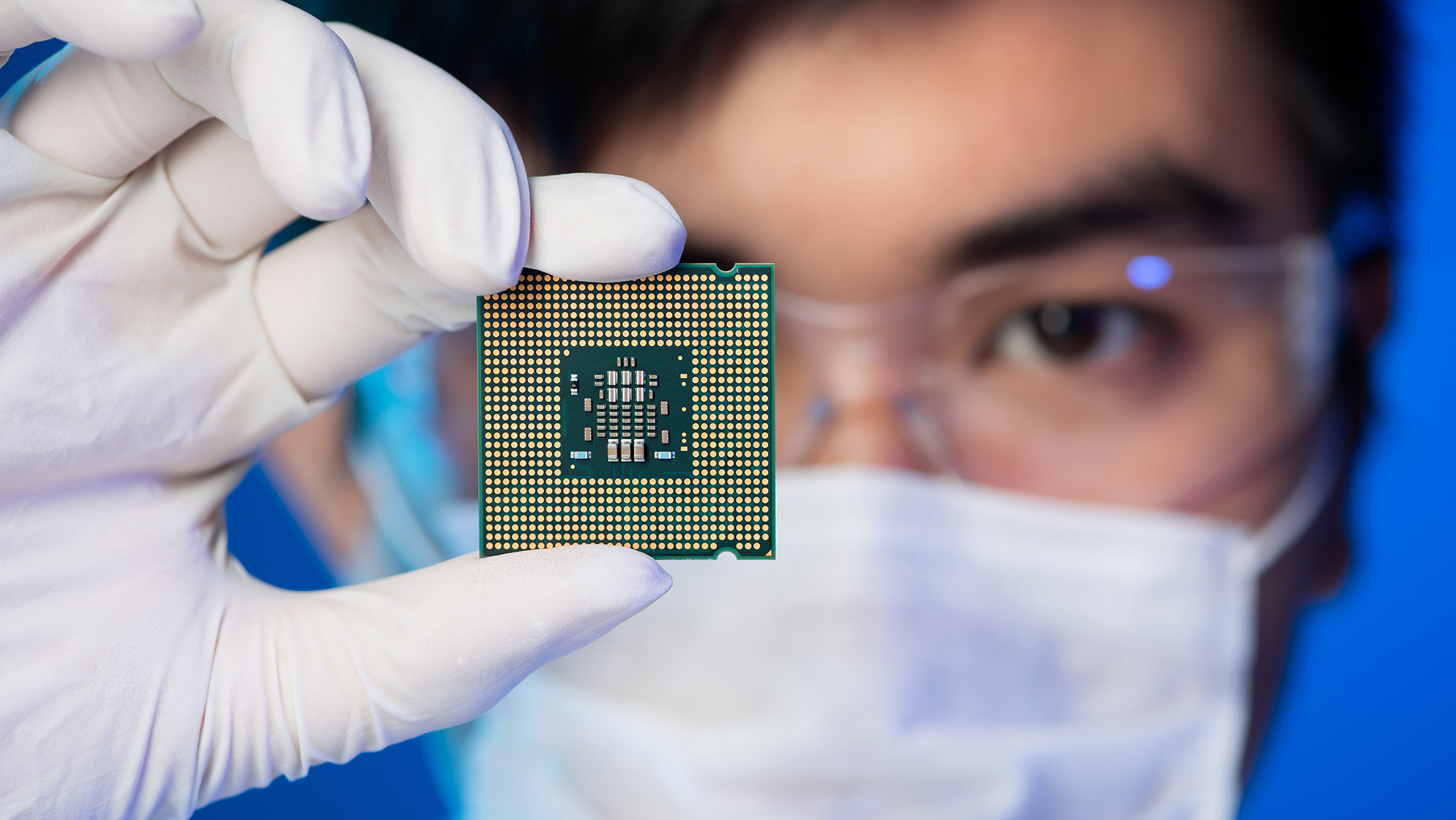 A person wearing lab safety attire holds a thin piece of square-shaped computer circuitry in a gloved hand