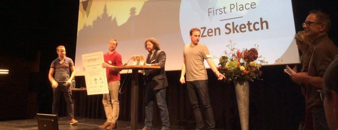 Six adults on stage at awards portion of competition. Man is standing behind podium reading from iPad. Another man to the right of him in a red shirt is holding a white sign. She image being projected on the curtain behind them says: "First place: Zen Sketch." There is a large metal vase of flowers on stage.