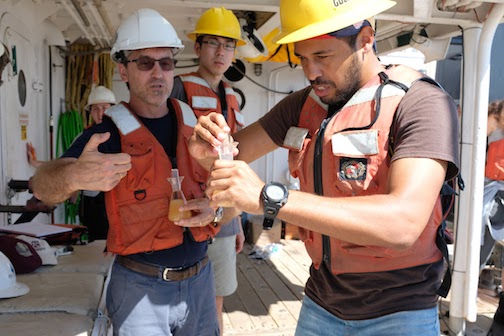 Three men in hardhats and orange safety vests. The man at the front is holding a tube of liquid. The man right behind him is holding a beaker of orange liquid. The other man is wearing glasses and is observing the actions of the first two men.