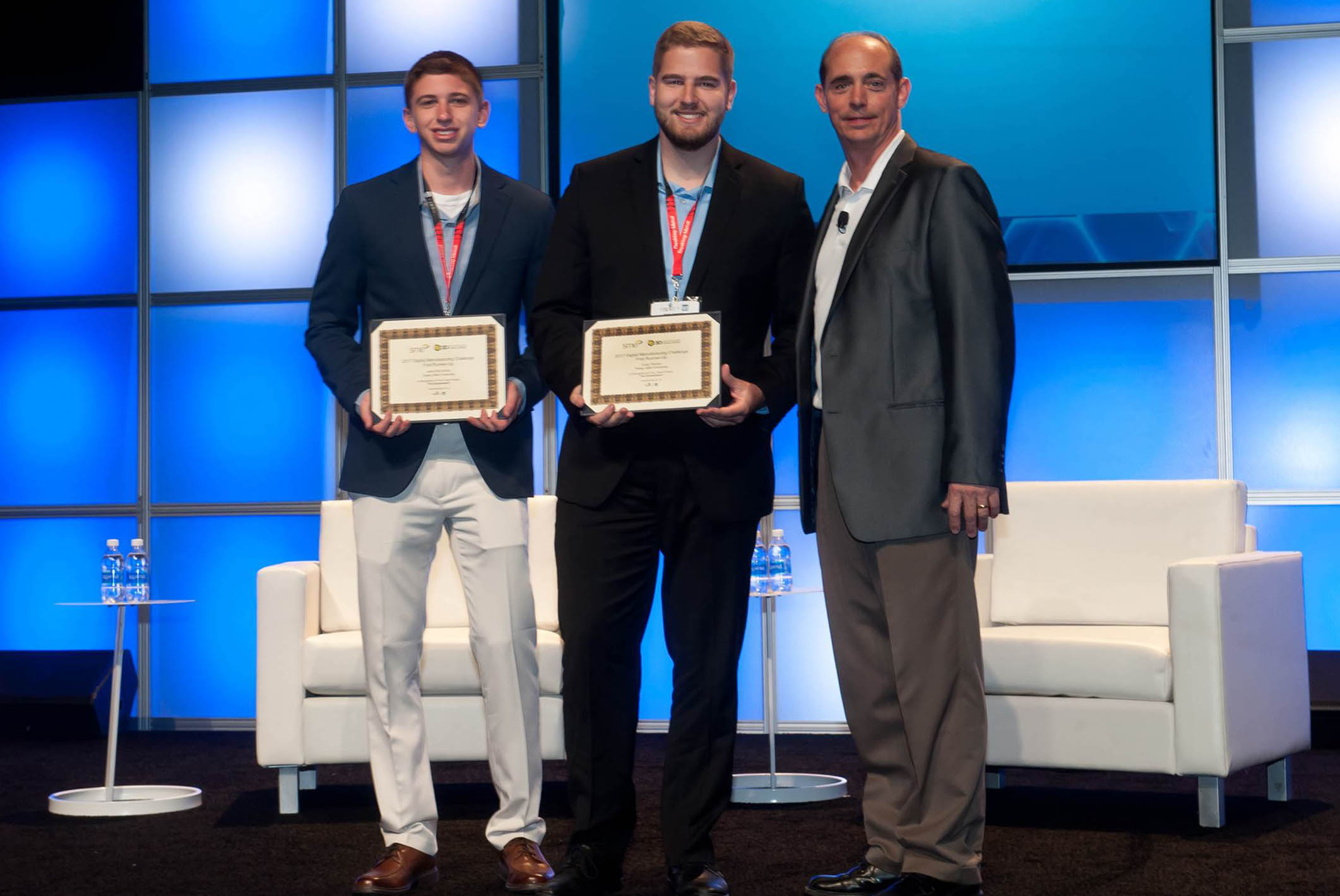 Three men on stage in business suits. Two are young men and they are holding certificates. There are two white plush chairs and an end table with water bottles on the top behind the three men. The wall behind the stage is several blue squares.