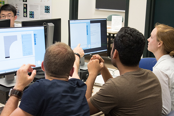 View of three students looking at two computer screens from behind. One student can be seen at another computer in the top lefthand corner.