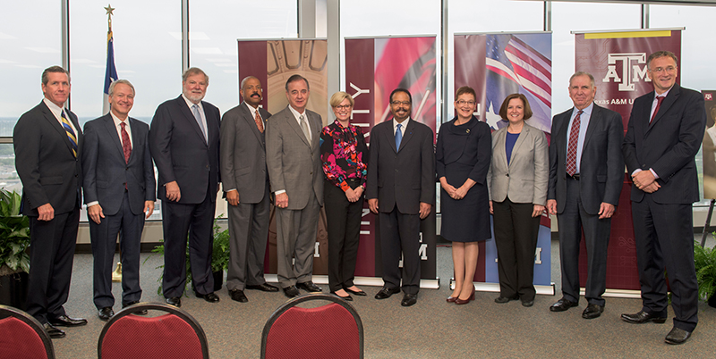 Eight men and three women in dark colored business clothing standing in room with wall of windows behind them. There are banners behind them with images and a maroon band with wording on the left side. Three chairs can be seen in the bottom left corner facing the group.