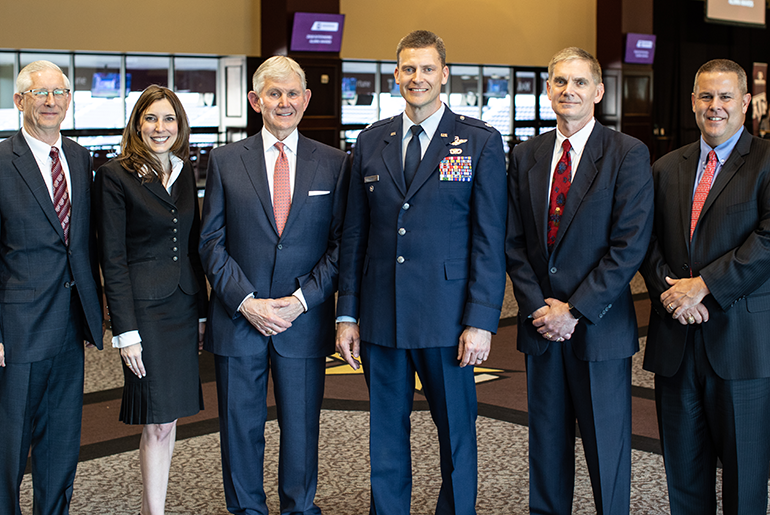 Group of five men and one woman is dark colored business attire in room where windows and two tv monitors can be seen in the background.