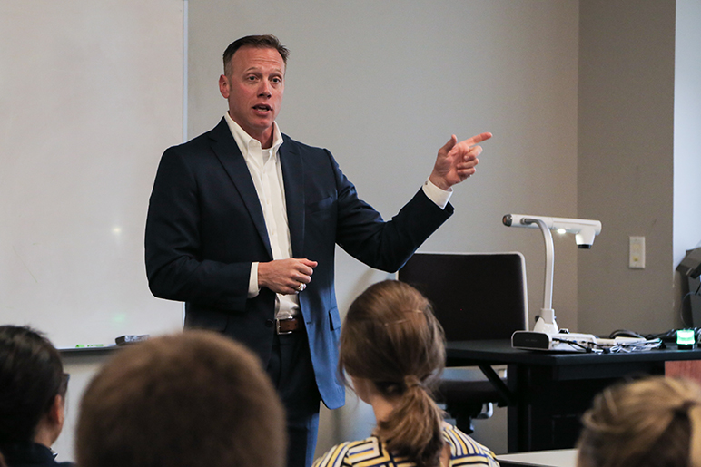 Man in dark blue blazer and white shirt and dark wash jeans standing in front of group of students in a classroom.