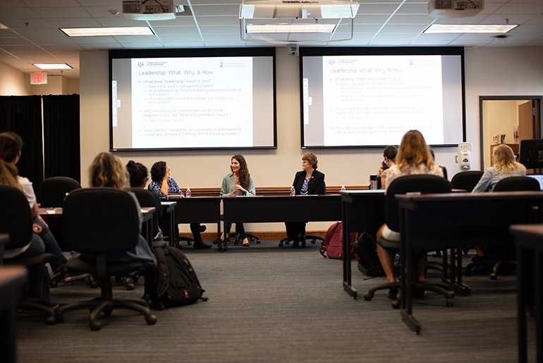 Four women are sitting at the front of a classroom at a long table with a powerpoint presentation being projected on two screens behind them. There is a small crowd of female students sitting at desks in rolling chairs listening to the women.