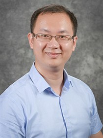 Man in light blue, short-sleeved button up shirt. He is shown from his shoulders up in a formal headshot. The background is a dark grey.