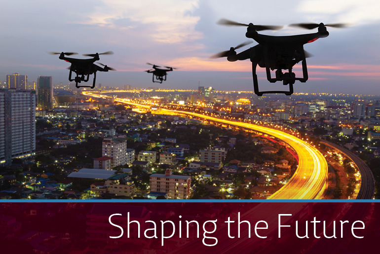 Aerial view of city landscape. Lights on highway are a yellow/gold blur to illustrate the speed at which traffic is traveling. In the sky are three drones. At the bottom right hand corner of the picture there is a maroon band with "Shaping the Future" on it in white font.