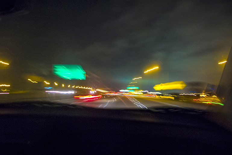 Camera is in drivers seat of car looking out at blurred scene of road, traffic lights and other cars during night.