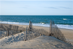 Beach with white sand and blue water. There is a wooden fence made up of thin, pieces of wood.