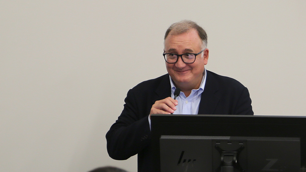 A man wearing glasses stands at a podium and speaks into a microphone