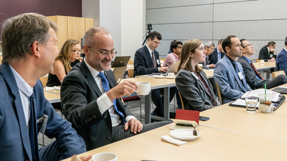 A group of 12 people seated in two rows of tables listens to an unpictured speaker. A gentleman wearing a gray suit is chuckling as he drinks from a cup of coffee.