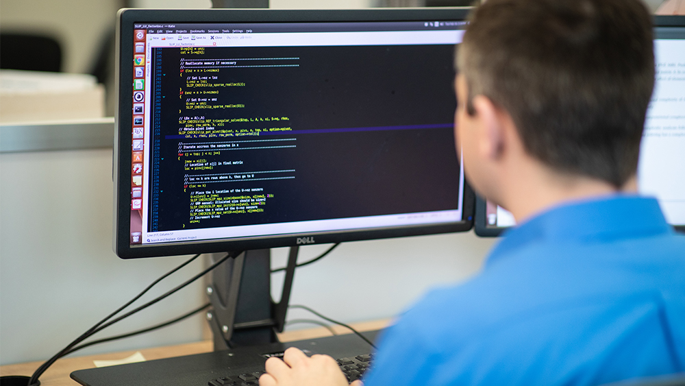 Student sitting at a desk looks at a computer screen showing computer coding. 