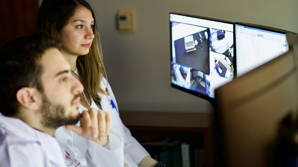 Two students in lab coats look at computer monitors during an experiment. 