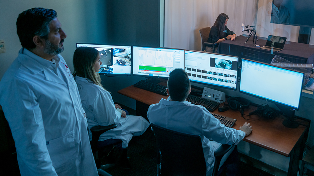 Professor observes students, who are looking at computer screens, who are conducting an experiment behind a one way mirror.