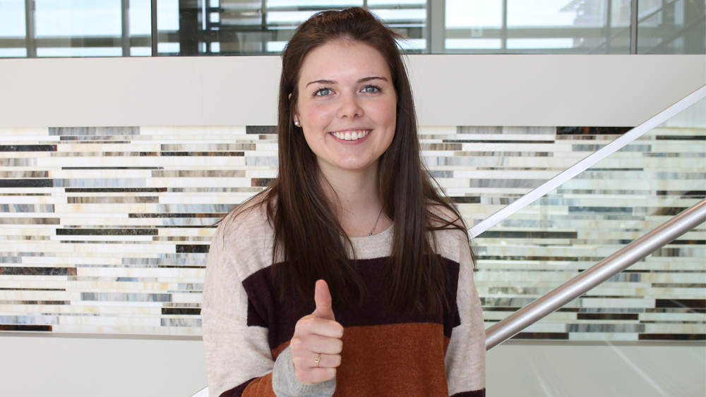 A girl with brown hair and gray eyes stands on stairs. She is giving a thumbs up, Gig 'em sign.