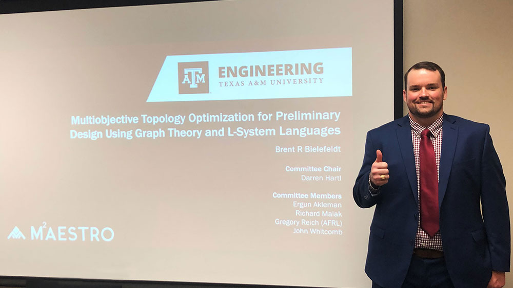 Brent Bielefeldt in a professional suit, posed next to a project screen showing the opening slide of his dissertation.