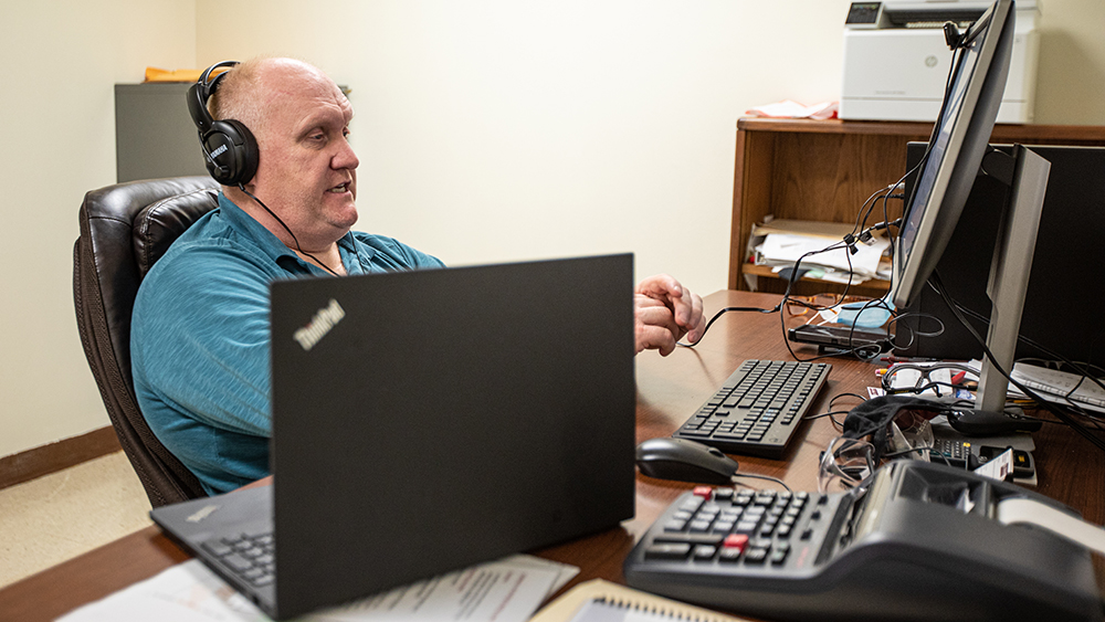 John Mahoney, lecturer, sitting at his  desk with headphones on working on a computer and other equipment.