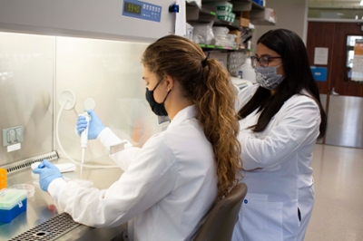 Female professor observes female student using pipette equipment in the lab.