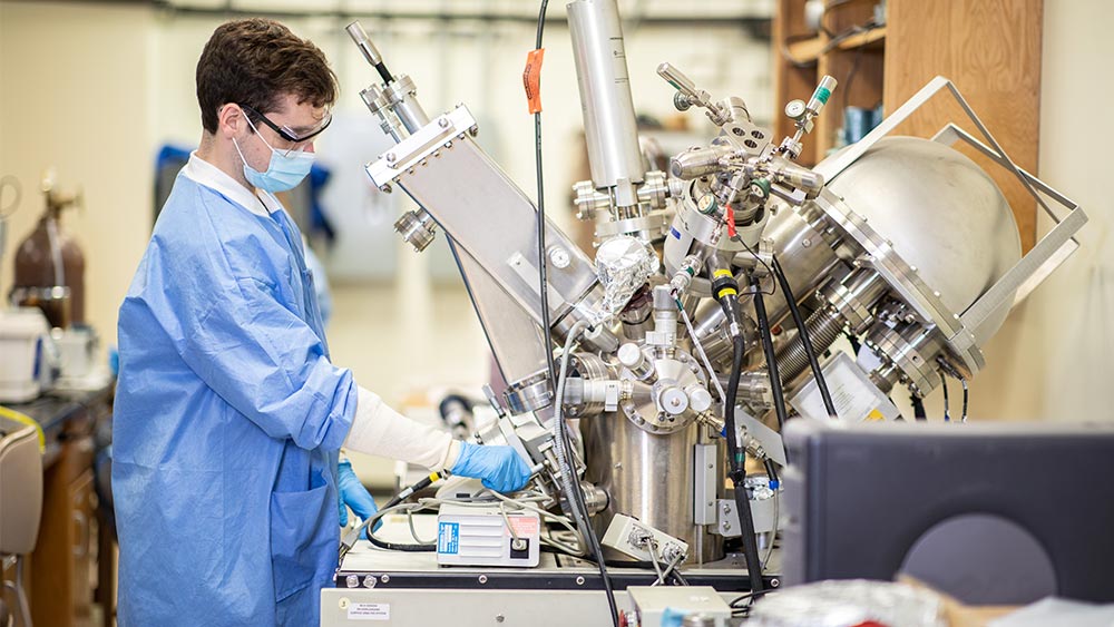 A student scientist dons protective laboratory gear while looking into a large microscope