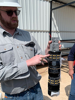 man wearing hard hat holding a two-foot long cylindrical mechanism in one hand while pointing to a feature on the cylinder with fingers of other hand