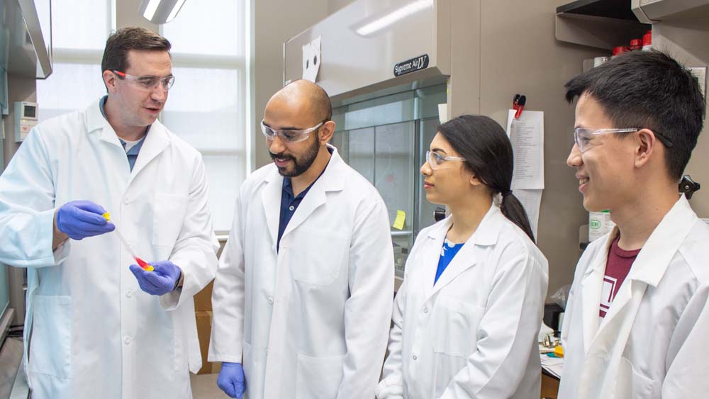 Faculty member holds a tube with a red liquid inside and shows it to a group of three students. All are wearing personal protective equipment.