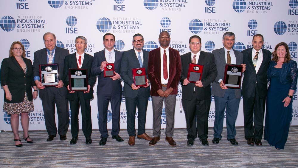 People stand in front of a white background. Many are holding awards. They are all smiling at the camera.