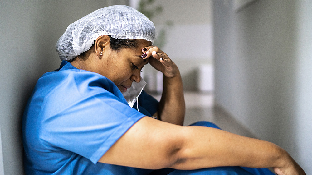 A Black female nurse sits on a dark hallway floor with back against wall, looking down with hand on her head, and a mask partially off her face, in a fatigued state