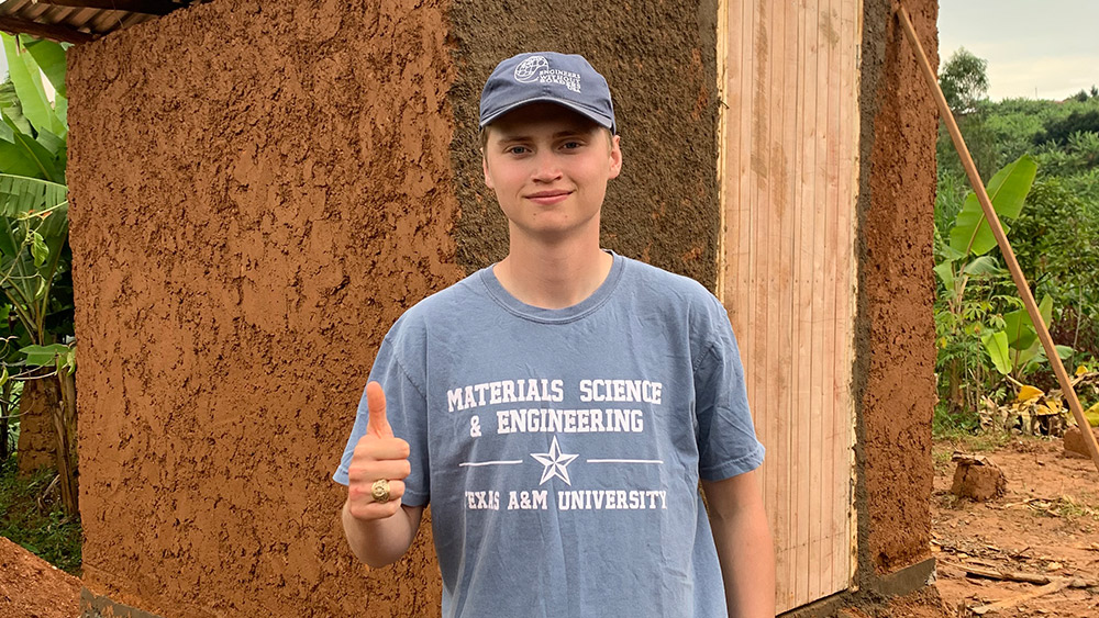 Eli Norris giving a thumbs up in front of a latrine made of concrete-like material with wooden door and metal roof