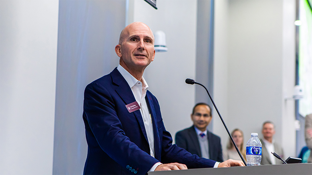 Ben Keating stands at a podium with a microphone. In the background, Dr. Bimal Nepal stands with attendees of the capstone showcase.