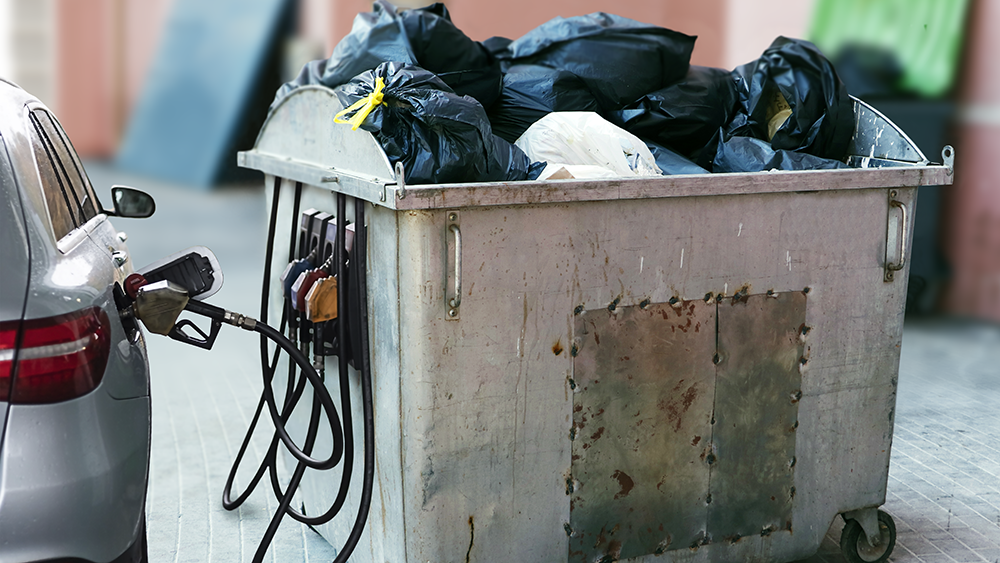 A car connected by gas pumps to a grey dumpster full of trash. The dumpster pumps are fueling the car with gas.