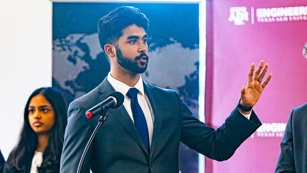 Ankit Shah speaks behind a microphone with one arm raised; Likitha Joneboina stands in the background.