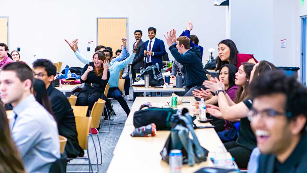 Group of students sitting at tables in a room. In the back, some stand and cheer while others applaud.