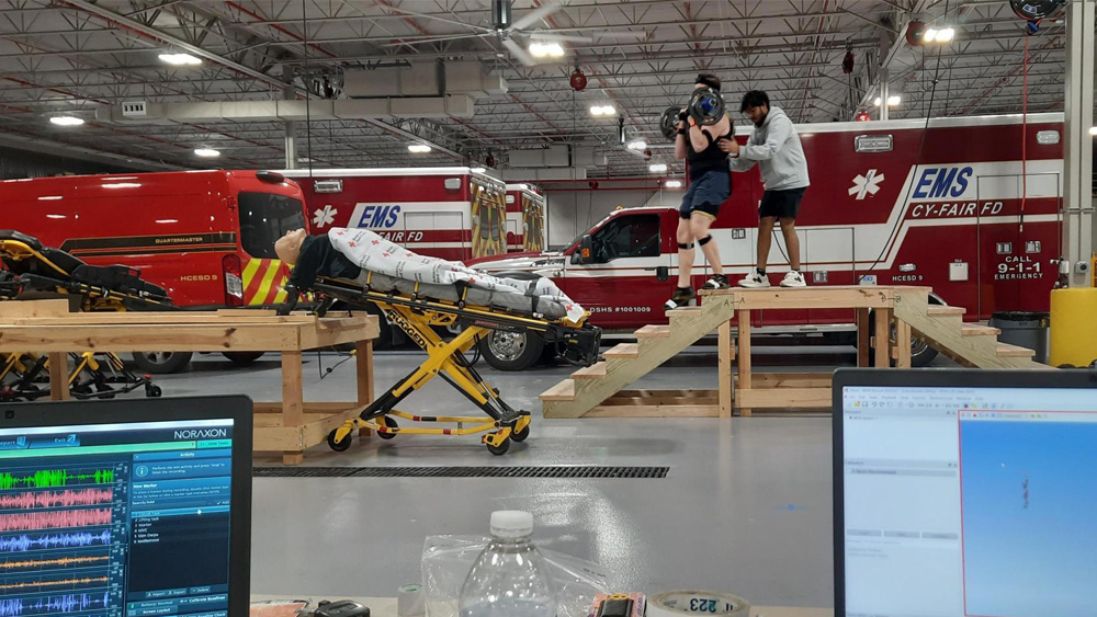 A student researcher stands with a medical technician at the top of a set of wooden stairs. The technician is wearing a backpack like device and holding weights. Computer screens show how the technician is being monitored for biometrics.