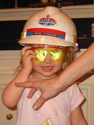 A mother’s hand reaches for a grinning two-year-old girl wearing a large dirty hard hat and plastic safety glasses.