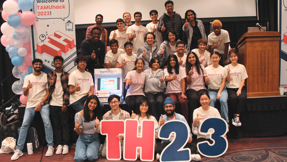 A group of TAMUhack students sitting on and in front of a stage doing a thumbs up.