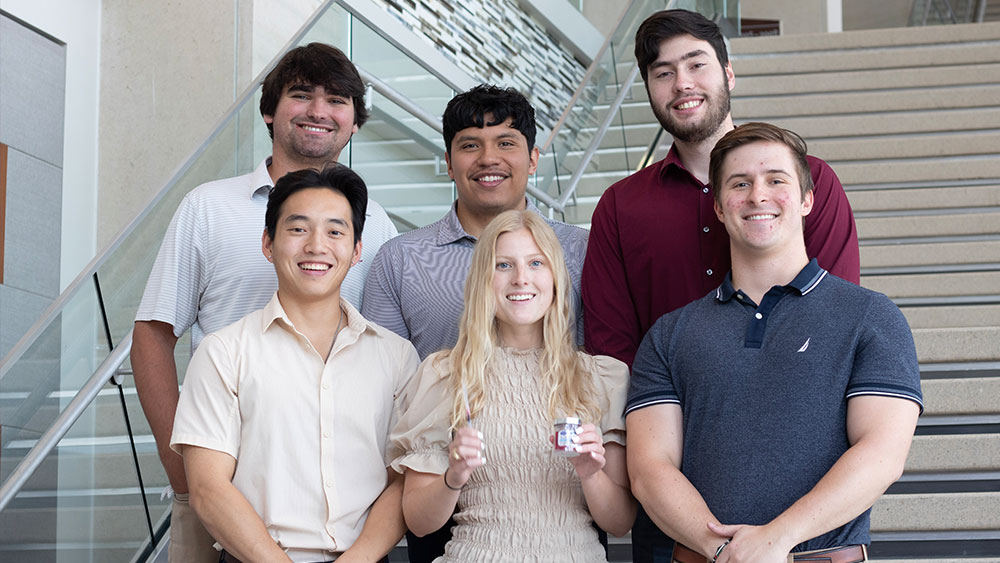 Five male and one female student stand on a staircase, smiling at the camera. The female student holds a vial and needle.