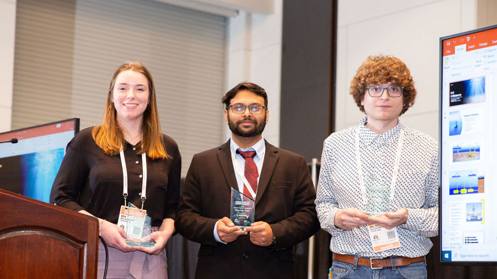 Three Texas A&M University students hold glass awards while they stand on a stage by a podium.