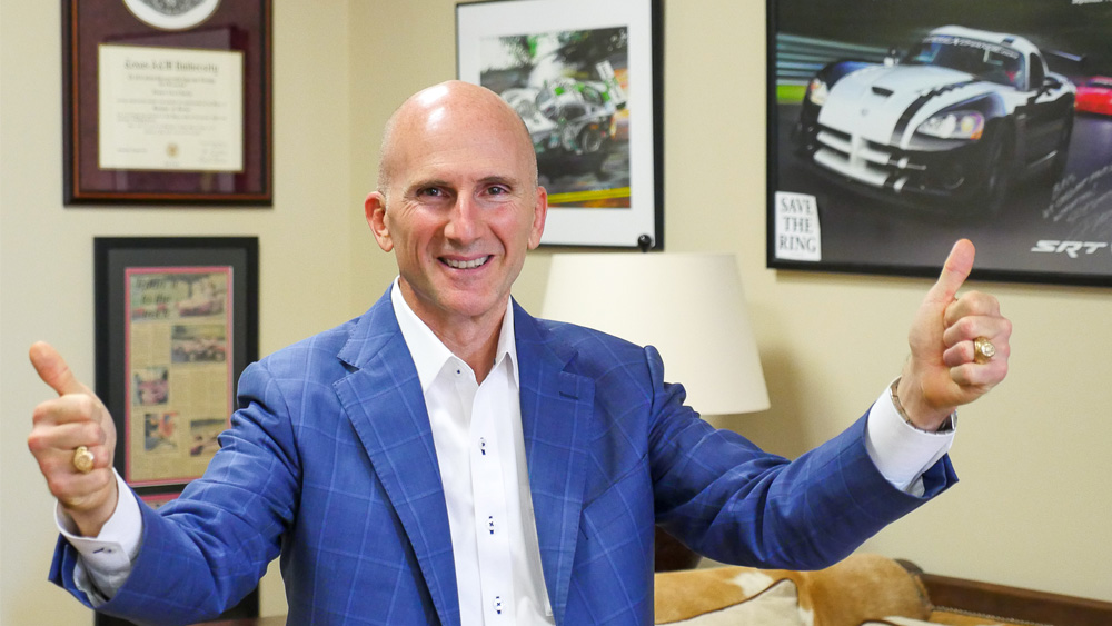 Ben Keating stands smiling at the camera, giving two thumbs up, showing two large gold rings.